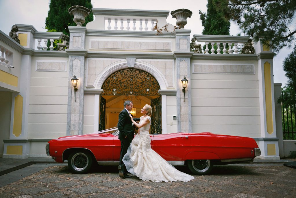 Bride and groom standing in front of red wedding car at the front doors at Illyria House in Pretoria
