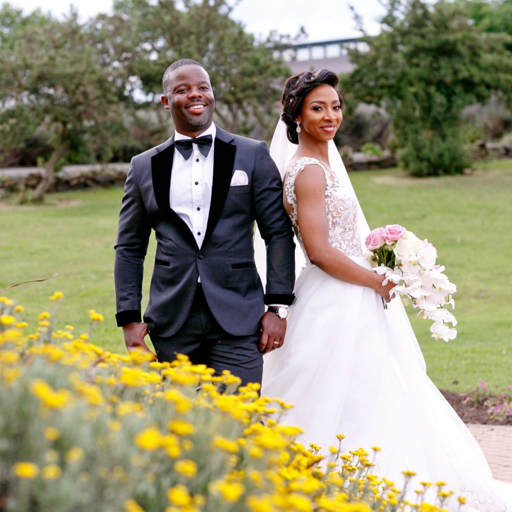 bride and groom posing in front of pretty yellow flowers at Bridesmaids in pretty pink dresses walking past Brahman Hills
